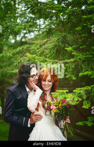 Deux jeunes mariés heureux, souriant et à proximité d'arbres verts. Cheveux rouge mariée. Mariage soirée d'été. Banque D'Images