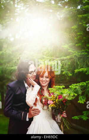 Deux jeunes mariés heureux, souriant et à proximité d'arbres verts. Cheveux rouge mariée. Mariage soirée d'été. Banque D'Images