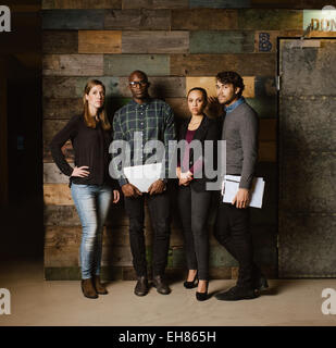 Longueur totale de l'image de l'équipe des Créatifs multiraciale qui pose pour appareil photo de bureau. Les jeunes gens d'affaires se tenant ensemble contre un wo Banque D'Images