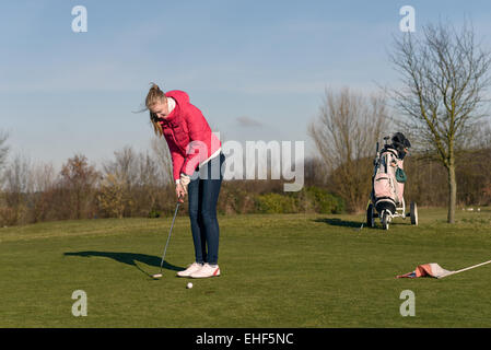 Woman alignement d'un putt sur le green avec son chariot de golf et clubs visible derrière sur un ciel bleu ensoleillé jour Banque D'Images