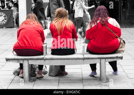 Trois jeunes femmes de différentes tailles wearing red tops assis sur un banc avec les piétons marcher passé en monochrome à Bristol Banque D'Images