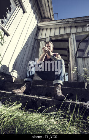 Un jeune homme assis à l'extérieur d'une maison abandonnée et alléger une fumée. Banque D'Images