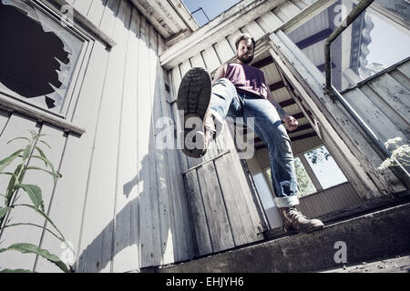 Un jeune homme de coups de pied et de fumer à l'extérieur d'une maison abandonnée. Banque D'Images