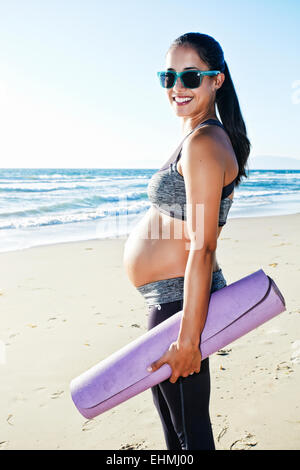 Pregnant Hispanic woman carrying yoga mat on beach Banque D'Images