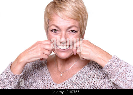 Belle femme d'âge moyen avec de courts cheveux blonds cherche à renverser les signes du vieillissement en tirant sur ses joues avec ses mains Banque D'Images