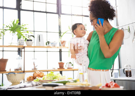 Mère tenant son bébé fille, alors qu'à l'aide de mobile phone in kitchen Banque D'Images