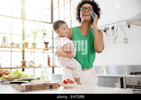 Mère tenant son bébé fille, alors qu'à l'aide de mobile phone in kitchen Banque D'Images