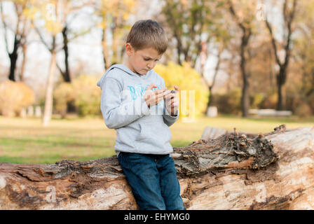 Boy leaning against tree trunk jouant sur appareil mobile dans le parc Banque D'Images