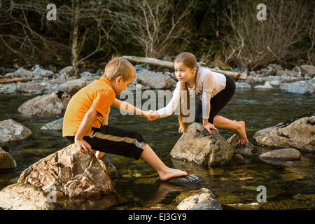 Neuf ans, fille d'encourager son frère de sept ans sur la façon d'avancer à travers les rochers dans la rivière peu profonde Banque D'Images