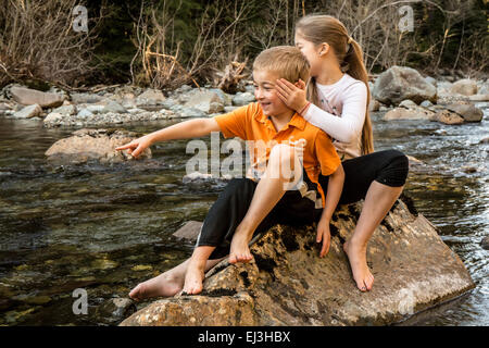 Neuf ans, fille de manière ludique couvrant les oreilles de son frère de sept ans, assis sur un rocher dans une rivière peu profonde Banque D'Images