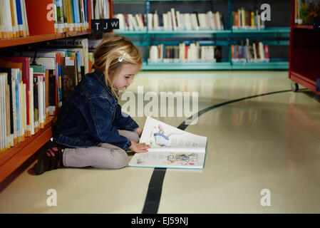 Caucasien enfant petite fille blonde à la recherche, la lecture et le choix des livres pour enfants sur l'étagère de bibliothèque publique Banque D'Images