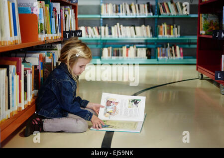 Caucasien enfant petite fille blonde à la recherche, la lecture et le choix des livres pour enfants sur l'étagère de bibliothèque publique Banque D'Images