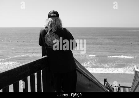 Accès à la plage Grandview à Encinitas, Californie, surfeur plus âgé avec casquette de balle et chemise avec légende Surf Dog, en regardant le surf Banque D'Images