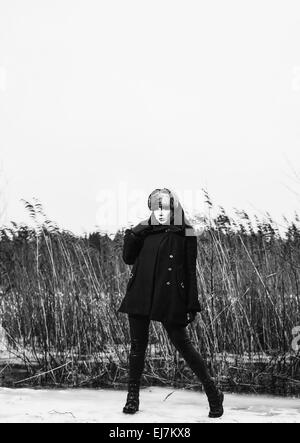 Fashion femme portant un manteau d'hiver et bonnet de fourrure et elle posant devant les roseaux, temps de pluie froide, pleine longueur noir et whi Banque D'Images