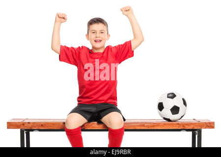 Junior joyeux joueur de football en chemise rouge gesturing bonheur assis sur banc en bois isolé sur fond blanc Banque D'Images