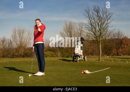 Jeune femme golfeur en jeans et blouson tenant un putter tout en donnant un coup de golf avec voiturette sur le côté. Banque D'Images