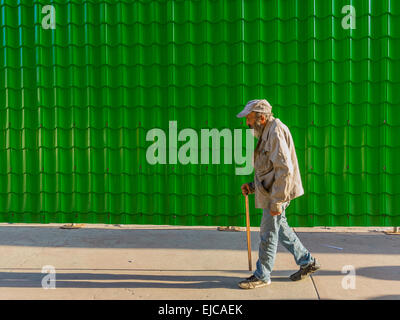 Un mâle cubain Senior citizen marche dans la lumière de fin d'après-midi avec une canne, le port d'un type baseball hat sur un fond vert. Banque D'Images