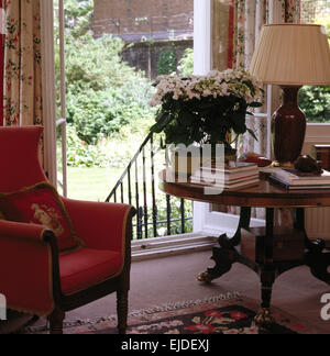 Fauteuil rouge à côté de table antique circulaire dans années 90 salon avec portes françaises s'ouvrent sur le jardin Banque D'Images