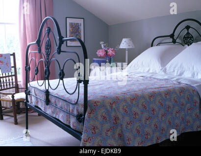 Couverture à motifs de roses et blancs ornés de coussins lit en fer forgé noir, gris pâle, dans une chambre mansardée avec rideaux rose Banque D'Images