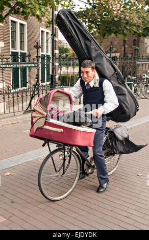 Bicyclist riding avec un violoncelle sur le dos et un panier avec son chien à Amsterdam. Banque D'Images