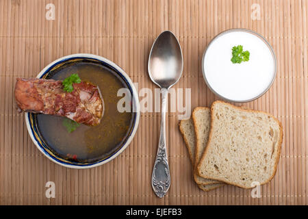 Soupe de légumes avec de la viande fumée, pain, crème et cuillère en argent sur nappe en vue de dessus. Banque D'Images