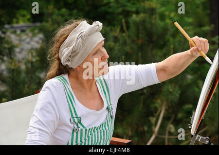 Senior woman painting à l'extérieur. Elle siège dans une toile chaise devant le chevalet. La journée est couvert en donnant une lumière douce Banque D'Images
