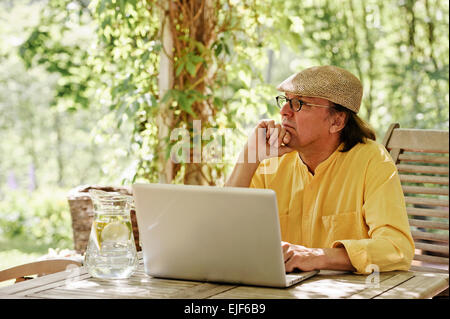 Hauts homme est assis à l'extérieur à une table en bois sous une pergola et travaille sur un ordinateur portable. C'est l'été et il y a un arrière-plan Banque D'Images