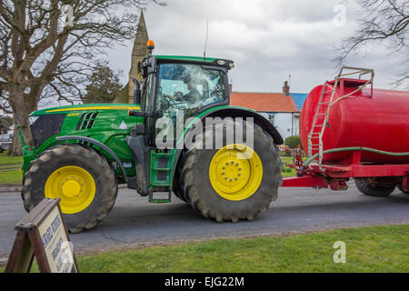 Un John Deere 6210R tracteur agricole de tracter une remorque réservoir à travers un village de County Durham England UK Banque D'Images