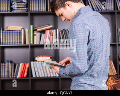 Jeune homme à la recherche des informations dans un livre qu'il a prises à partir d'une étagère dans la bibliothèque comme il fait de la recherche pour un projet Banque D'Images