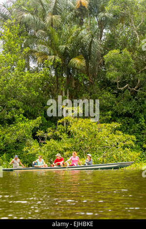 Cinq personnes sont assises dans un long canot étroit sur une rivière calme et verdâtre entourée d'une végétation tropicale dense. Le groupe comprend trois femmes et deux hommes, portant des vêtements de plein air décontractés et des chapeaux Banque D'Images