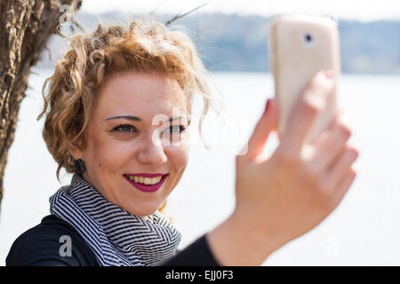 Jolie jeune femme blonde aux cheveux bouclés en tenant le téléphone avec selfies en extérieur.Elle sourit alors qu'elle communique avec elle Banque D'Images