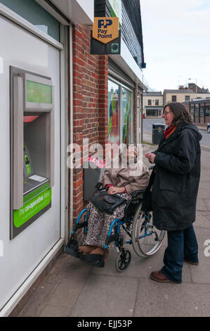 Une vieille dame vieille de 90 ans dans un fauteuil roulant a aidé à retirer de l'argent à un distributeur automatique distributeur automatique de par sa garde ou assistant. Banque D'Images