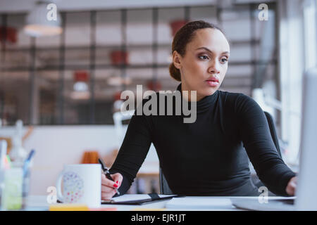 Image de jeune femme africaine nouvelle affectation de travail d'entreprise. La femme assise à son bureau à l'aide d'ordinateur portable et l'écriture remarque Banque D'Images