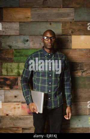 Portrait of young African man with a laptop looking at camera en position debout contre un mur en bois à l'office. Banque D'Images
