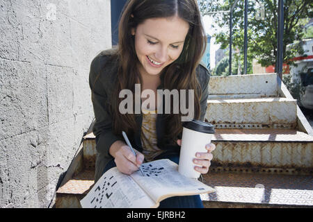 Caucasian woman doing crossword et boire du café sur les marches Banque D'Images