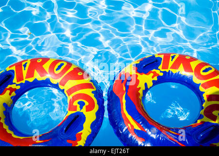 Deux gros anneaux en plastique gonflable bleu avec 'tropic' imprimé en rouge et jaune, flottant dans une piscine. Banque D'Images