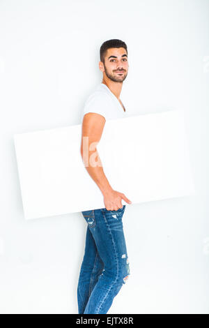 Jeune homme séduisant habillé décontracté et souriant montrant un panneau blanc sur fond blanc Banque D'Images