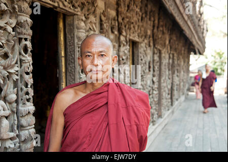 Portrait d'un moine bouddhiste en face d'un ancien mur de temple en bois sculpté avec un moine marchant en arrière-plan le Myanmar Banque D'Images