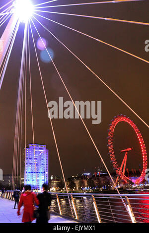 London England UK. Le 02 avril 2015. Après la tombée de la nuit sur la rive sud du London Eye est éclairé avec les feux rouges de l'actuel sponsor Coca-Cola et le bâtiment Shell devienne bleu. Vue depuis le pont piétonnier du jubilé sur la Tamise. Banque D'Images