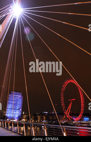 London England UK. Le 02 avril 2015. Après la tombée de la nuit sur la rive sud du London Eye est éclairé avec les feux rouges de l'actuel sponsor Coca-Cola et le bâtiment Shell devienne bleu. Vue depuis le pont piétonnier du jubilé sur la Tamise. Banque D'Images