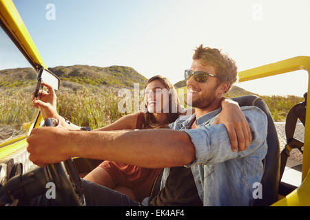 Homme conduisant beach buggy avec femme en tenant sur son selfies téléphone intelligent. Couple having fun on road trip sur une journée d'été. Banque D'Images