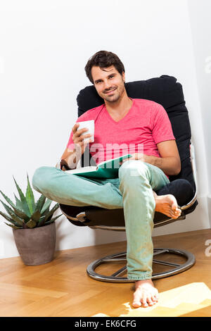 Jeune homme dans des vêtements décontractés assis sur chaise noir tout en lisant un livre et tenant un verre de boisson. Banque D'Images