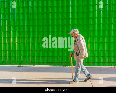 Un mâle Hispanic Senior citizen marche avec une canne à côté d'un mur fait de vert vif de tuiles produisant une répétition de la forme. Banque D'Images