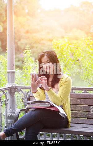 Belle jeune femme à l'université avec des livres et des dossiers sur les genoux, appuyant sur les boutons sur son téléphone, assis sur le banc en bois. Banque D'Images