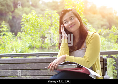 Belle jeune femme à l'université avec des livres et des dossiers sur les genoux, en utilisant son téléphone, assis sur le banc en bois dans un parc. Banque D'Images