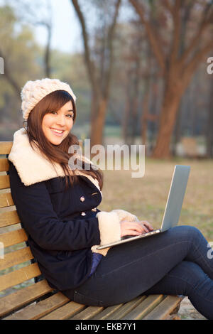 Jeune fille assise avec un ordinateur portable sur un banc dans un parc à l'hiver looking at camera Banque D'Images