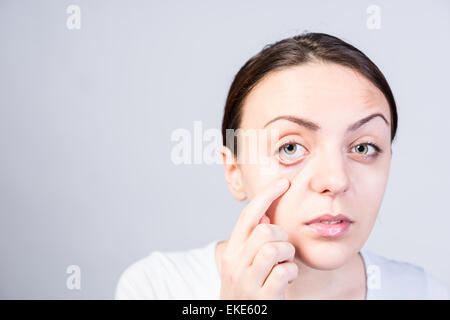 Close up jolie jeune femme tirant sa paupière inférieure en regardant la caméra sur un mur gris très clair avec l'exemplaire de l'espace. Banque D'Images