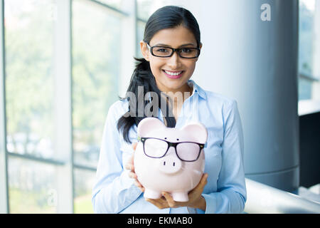 Closeup portrait heureux, smiling business woman, holding pink piggy bank, portant des lunettes noires grand bureau intérieur isolé Banque D'Images