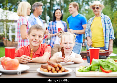 Frères et sœurs mignon avec saucisses de manger en plein air avec leurs parents sur l'arrière-plan Banque D'Images