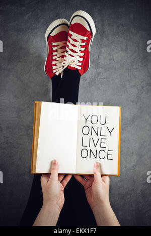 On ne vit qu'une fois ou de YOLO Concept with Young Woman Reading Book avec ses pieds soulevés en l'air Banque D'Images